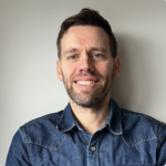 A man with short brown hair and a beard smiles at the camera. He is wearing a blue denim shirt and is standing in front of a light gray wall.